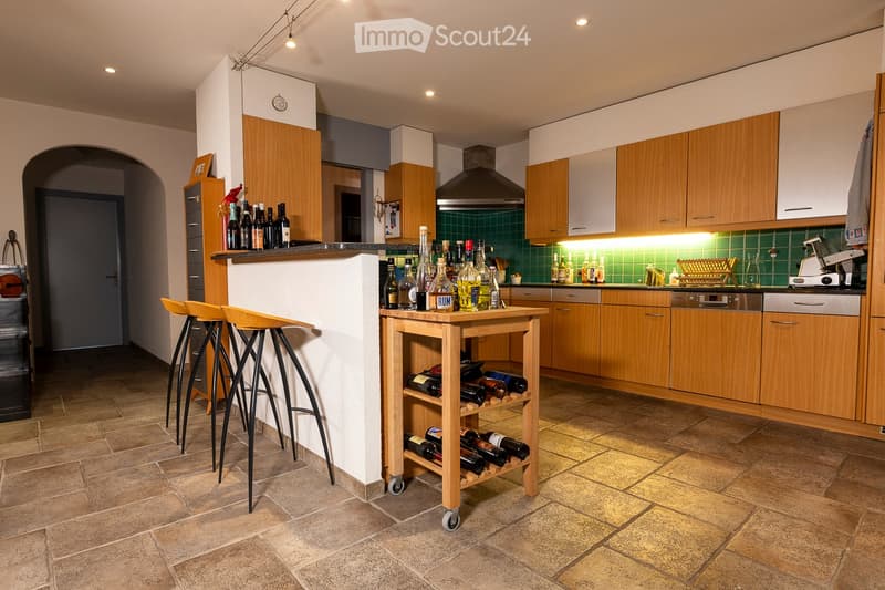 Modern kitchen with wooden cabinets, a central island, bar stools, and a wine rack. The kitchen features a tiled backsplash and a variety of bottles on the island and shelves.
