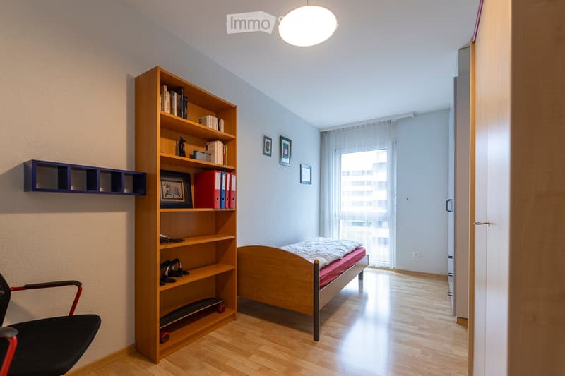 A modern, well-lit bedroom with a wooden bed, a large bookshelf filled with books and decorative items, a black chair, and framed pictures on the wall. The room features light wooden flooring and large windows with sheer curtains.