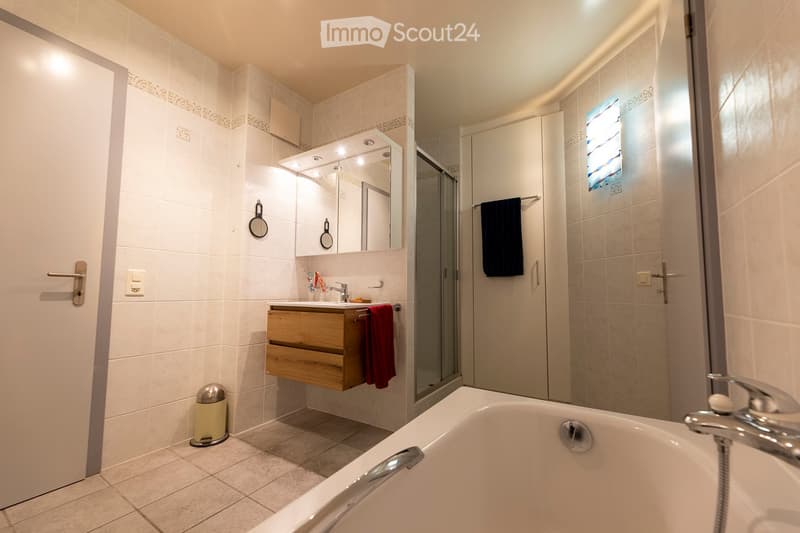 Modern bathroom with a bathtub, sink, mirror, and wooden vanity. Features tiled walls, a glass shower enclosure, and decorative towels.