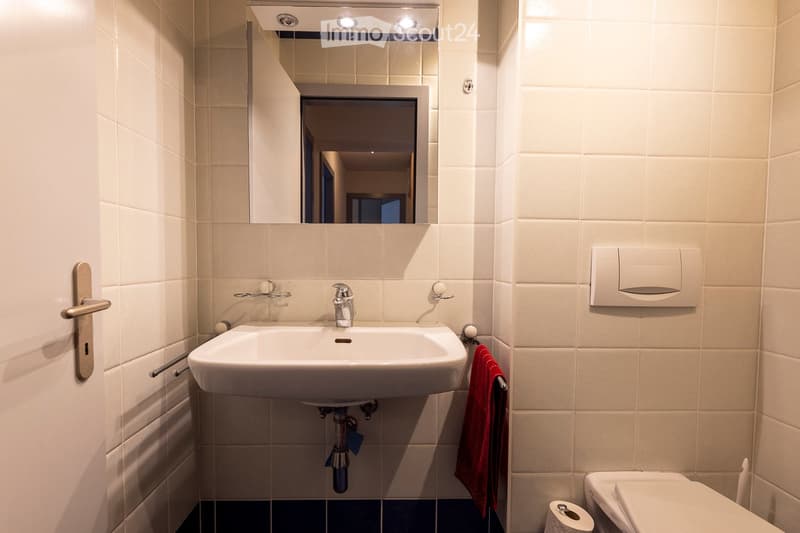 Modern bathroom with white tiles, a sink with a chrome faucet, a large mirror with built-in lighting, a toilet, and a red towel hanging on a rack.