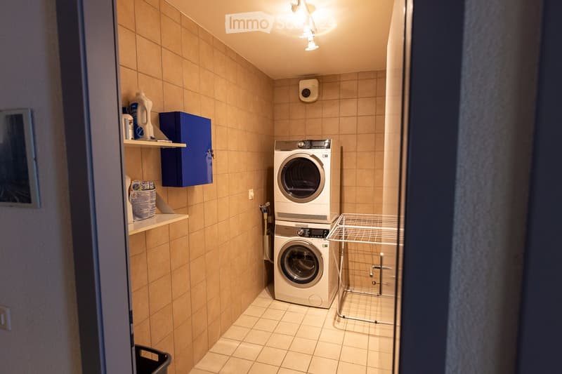A compact bathroom with tiled walls and floor, featuring a washing machine and dryer stacked vertically, a small shelf with cleaning supplies, and a blue cabinet.