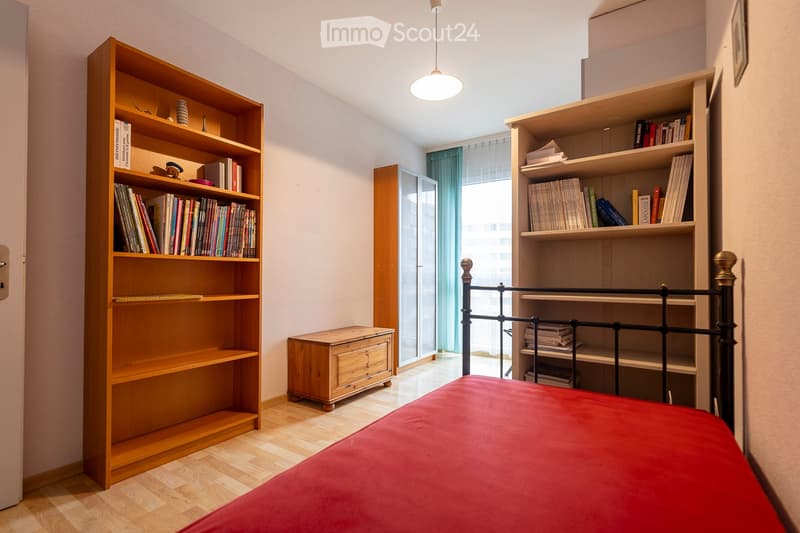 Cozy bedroom with wooden floor, red bedspread, two wooden bookshelves filled with books, a small wooden drawer, and a hanging lamp.
