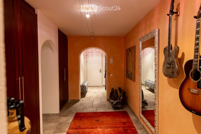 A narrow hallway with warm-toned walls, tiled flooring, and decorative elements including guitars, a mirror, and a red rug.