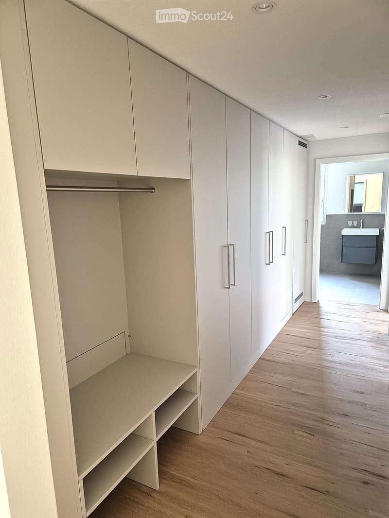Empty hallway with light-colored wooden flooring, built-in storage units with shelves and hanging rods, and a small adjacent bathroom visible at the end.