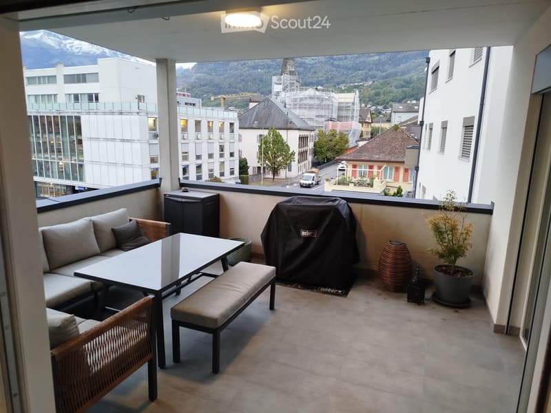 Balcony with a view of mountains and cityscape, furnished with a sofa, table, bench, chair, grill cover, vase, and potted plant.