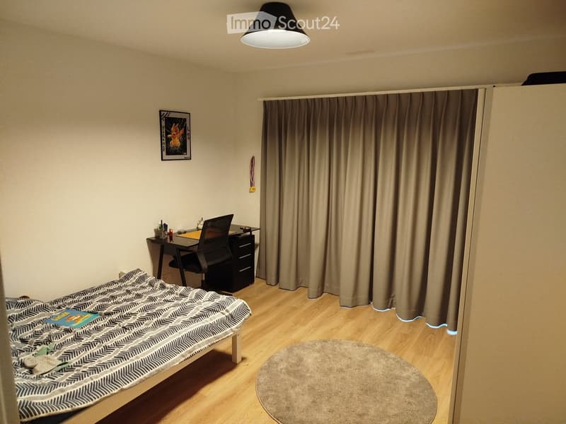 A small, furnished room with a bed, desk, chair, and a round rug. The room features wooden flooring, a large window with curtains, and a picture frame on the wall.