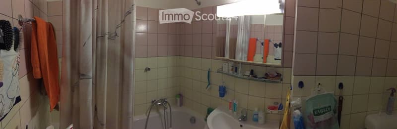 A small bathroom with a bathtub, sink, mirror, and various toiletries. The walls are tiled in a light pink color, and there is a shower curtain visible. A towel hangs on a rack, and a hairbrush is placed on the wall.