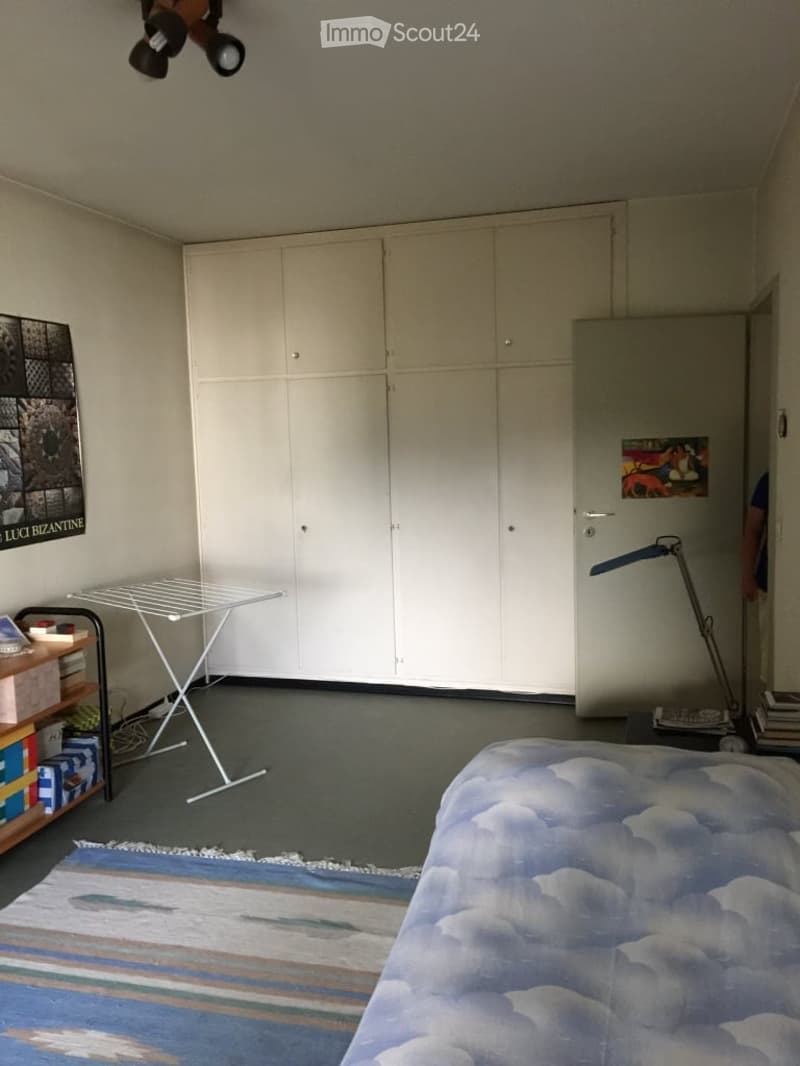 A tidy bedroom with a bed, large white wardrobe, small table, shelf, and a lamp. The room features a colorful rug and a picture frame on the wall.