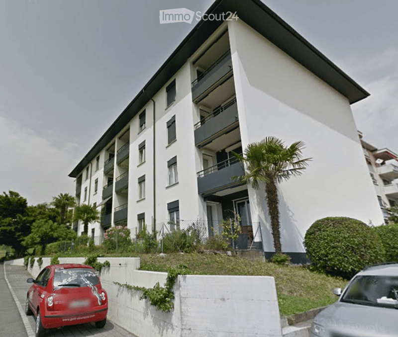 Modern multi-story apartment building with white facade, multiple balconies, and a raised ground floor. A palm tree and small garden area are visible in front of the building.