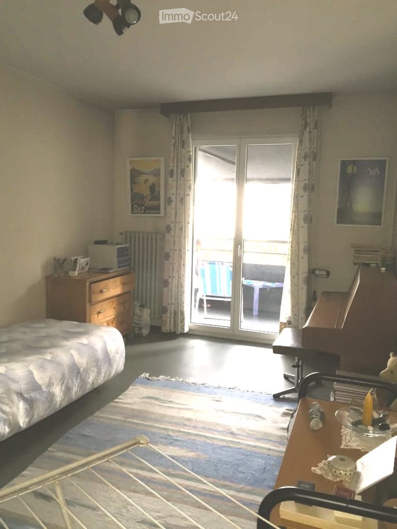 Cozy bedroom with a bed, nightstand, small table, chair, piano, and a sliding glass door leading to a balcony.