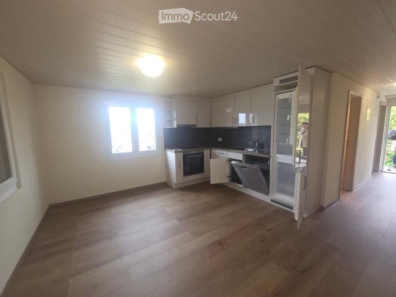 Modern kitchen with light wood flooring, white cabinets, and stainless steel appliances including a refrigerator, oven, and dishwasher. The space is well-lit with natural light from a large window.