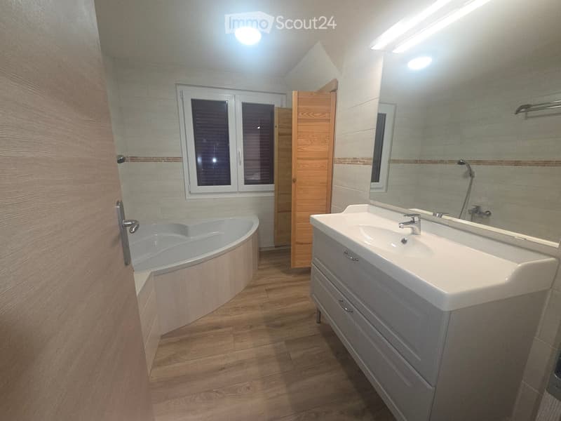 Modern bathroom with a wooden floor, a bathtub, a double sink with a cabinet, and a large mirror above the sink. The room features a window with blinds and a wooden door.