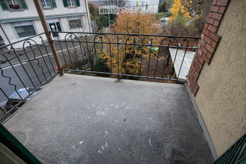 Balcony with metal railing, concrete floor, overlooking a street with autumn-colored trees and parked cars.