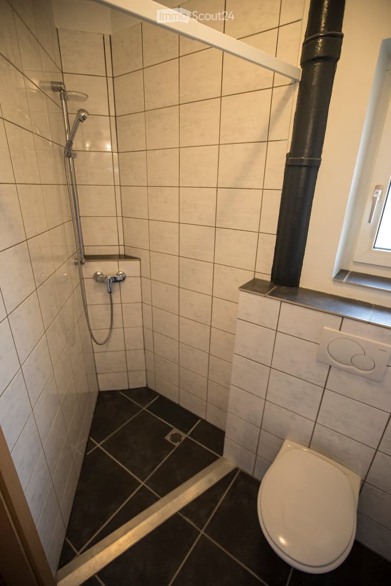 Modern bathroom with white tiled walls, dark tiled floor, toilet, shower, and window.