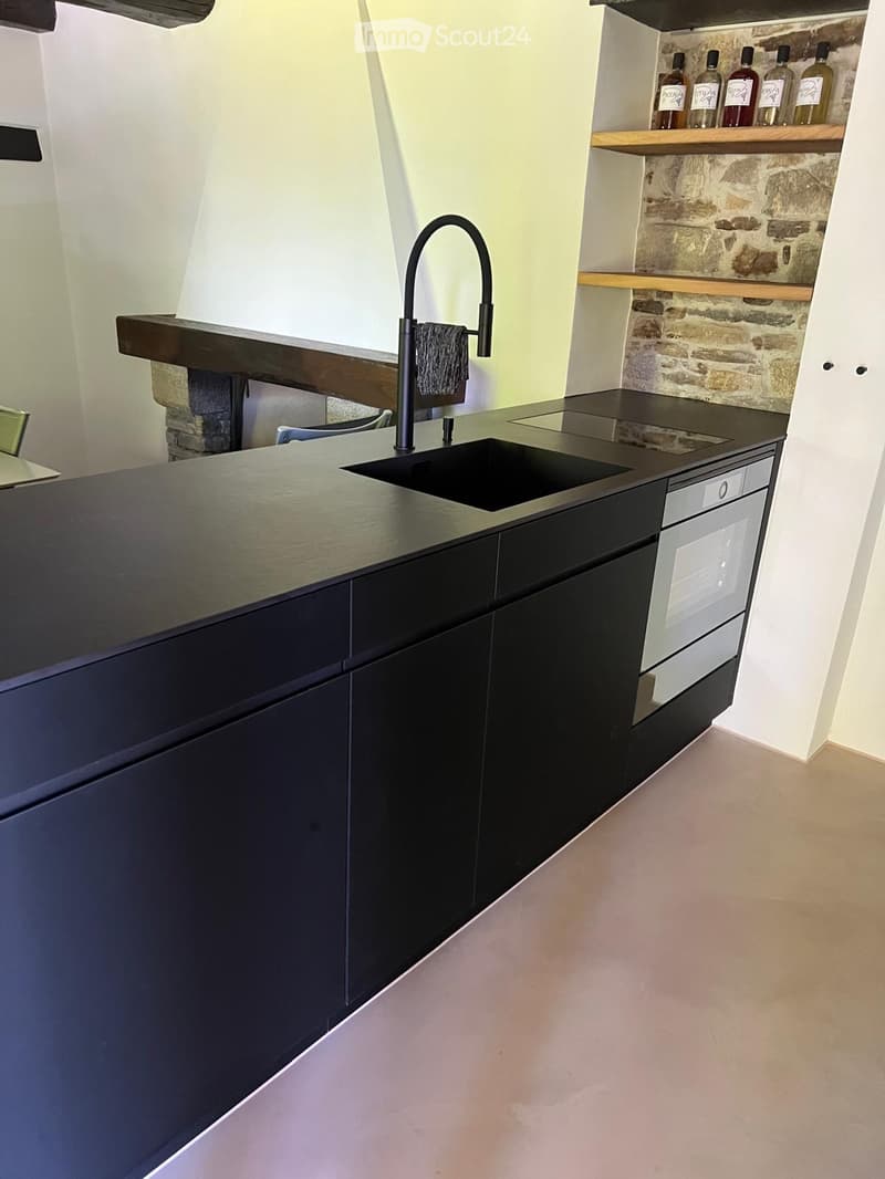 Modern kitchen with dark cabinets, a stainless steel sink, a sleek faucet, and a built-in oven. Shelves hold several bottles, and a stone wall adds texture.