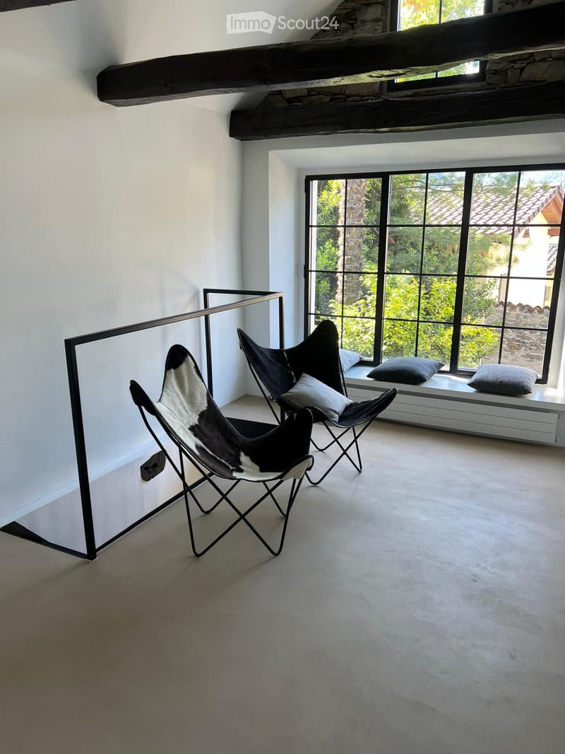 Modern, minimalist room with exposed wooden beams, large windows, and two black leather chairs with white cushions.