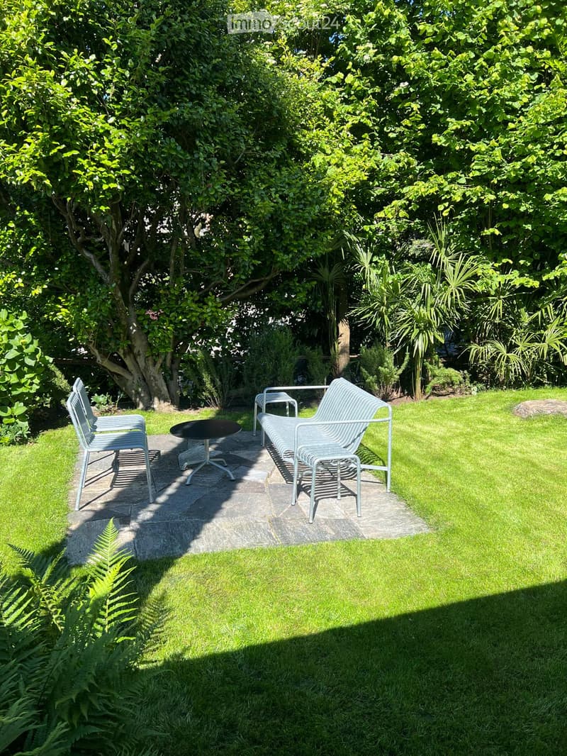 A well-maintained garden with a seating area featuring a bench, chairs, and a small table, surrounded by lush greenery and trees.