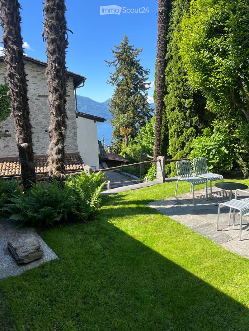 A well-maintained garden with a lawn, a few chairs and a small table, surrounded by tall trees and a view of a lake and mountains in the background.