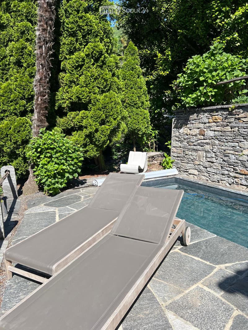 A serene outdoor area with a swimming pool, two sun loungers, and a white chair, surrounded by lush greenery and a stone wall.