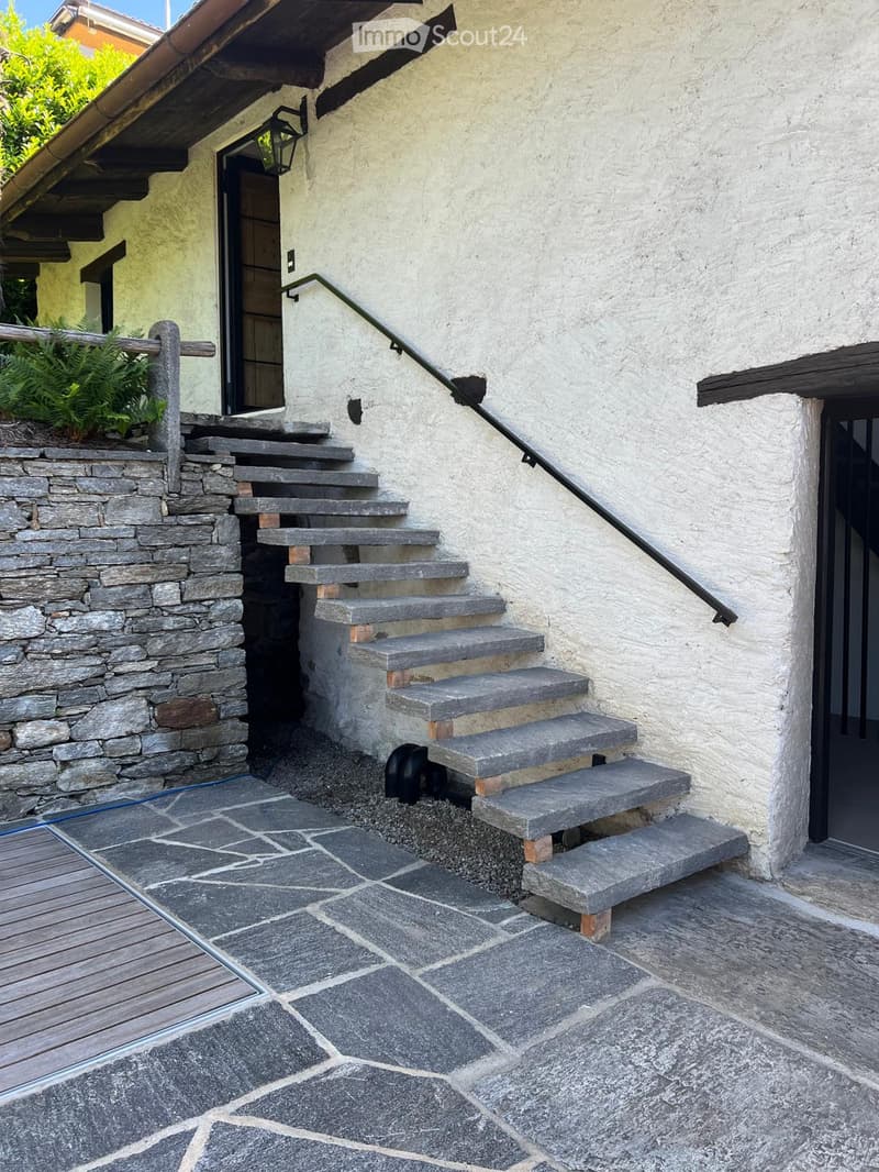 Stone steps leading up to a house entrance, with a metal railing and a small garden area on the left.