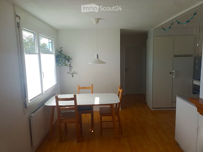 Bright dining area with a white table and four wooden chairs, large windows, and a modern kitchen area with cabinets and a refrigerator.