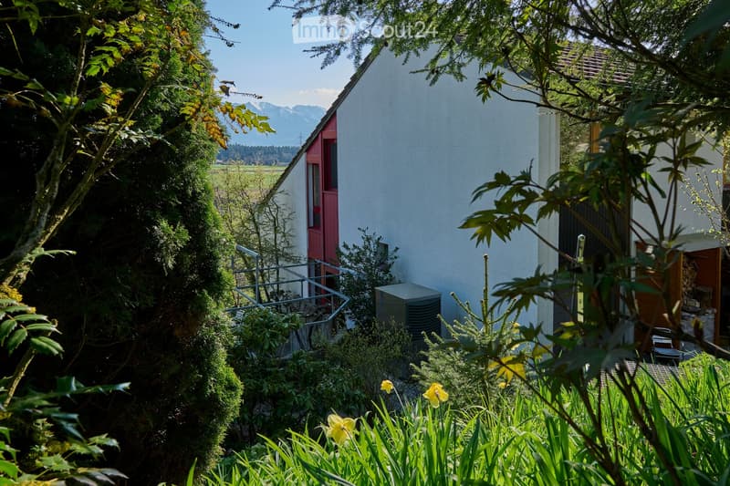 A serene garden area with a modern house in the background, surrounded by lush greenery and vibrant yellow flowers. The house features a white exterior with a red door and a visible air conditioning unit. The garden includes various plants and shrubs, wit