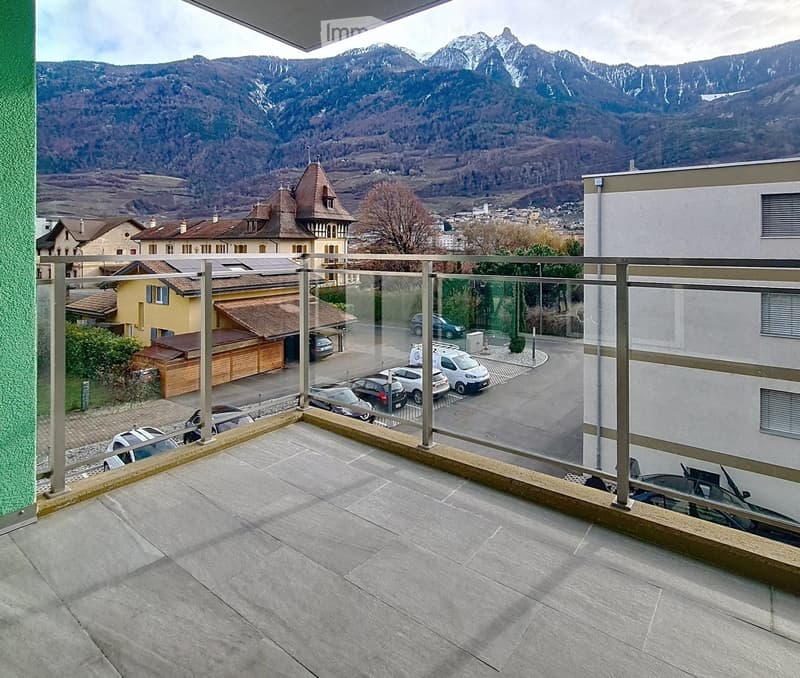 Balcony with a modern glass railing, overlooking a town and mountain range, with several parked cars visible below.