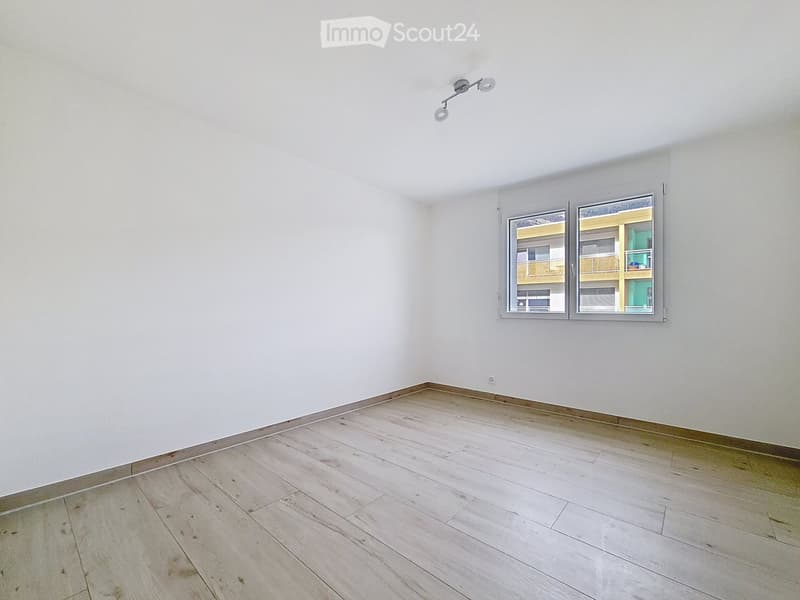 Empty room with light wooden flooring, white walls, and a window showing a view of neighboring buildings.
