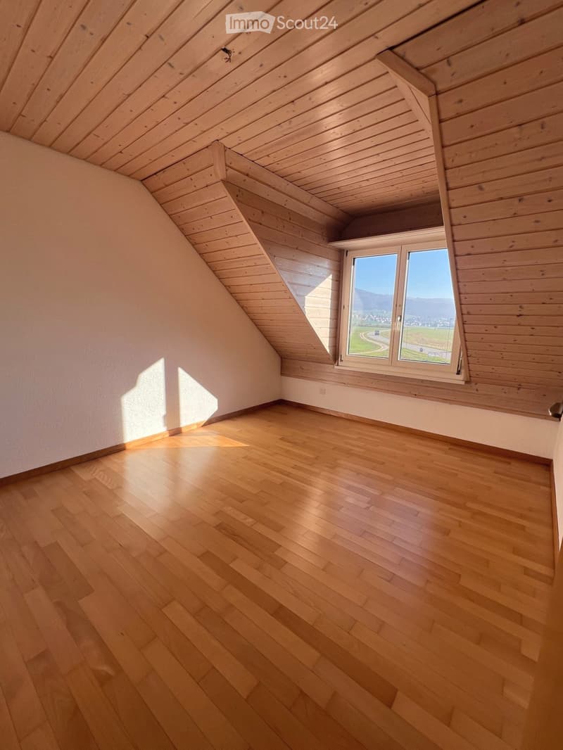 Empty attic room with wooden floors and slanted wooden ceilings, featuring a large window with a scenic view.