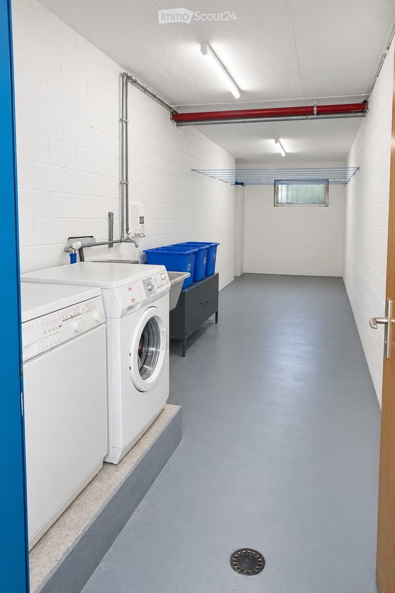 Clean, well-lit hallway with white brick walls, grey floor, and visible washing machine and sink area on the left side.