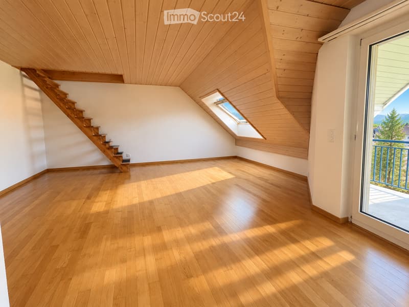 Bright, empty room with wooden flooring and slanted wooden ceiling. Features a staircase leading upwards, a skylight window, and a large glass door opening to a balcony with a view.