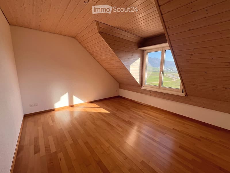 Empty attic room with wooden floors, slanted wooden ceiling, and large window offering a view of the landscape.