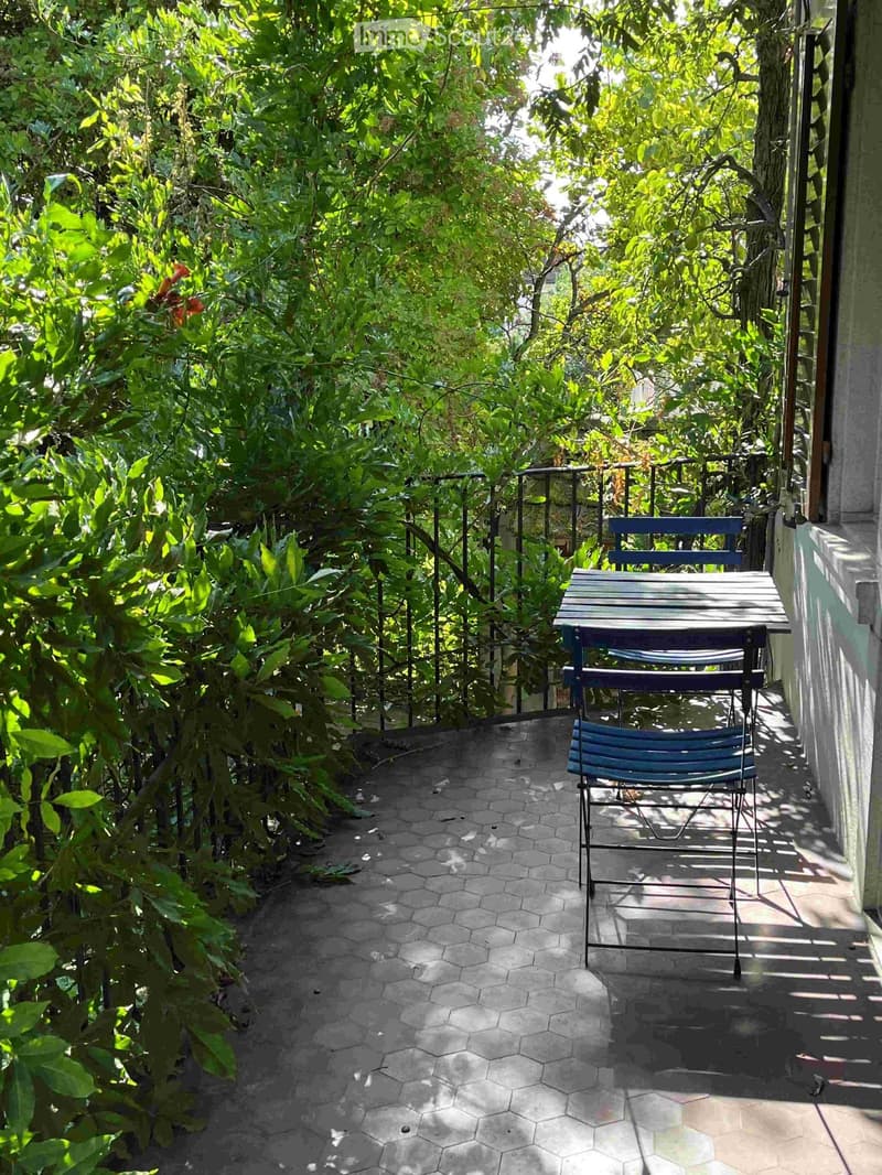 A small, enclosed balcony with a hexagonal tile floor, a table, and a chair surrounded by lush greenery and a metal railing.