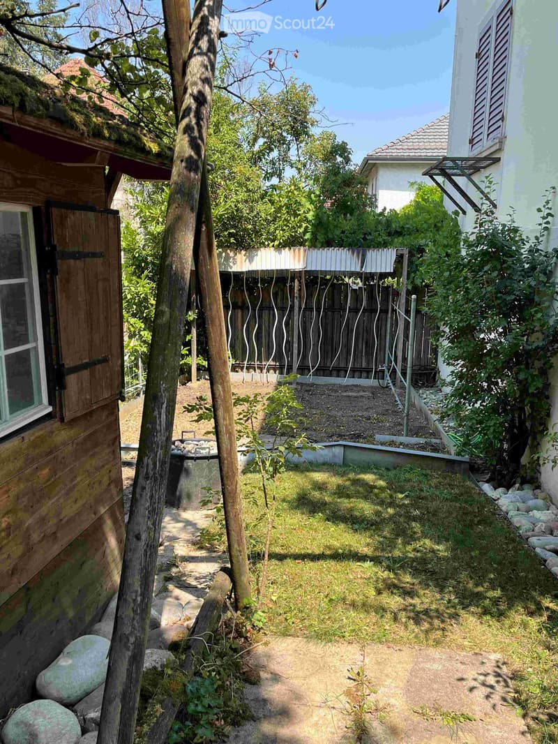 Garden area with a wooden fence, trees, rocks, and a small raised bed, surrounded by a house and another building.