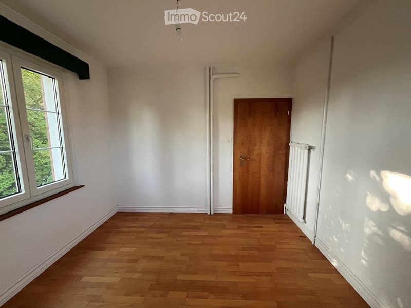 Empty room with wooden flooring, white walls, a window with a view of greenery, a wooden door, and a radiator.