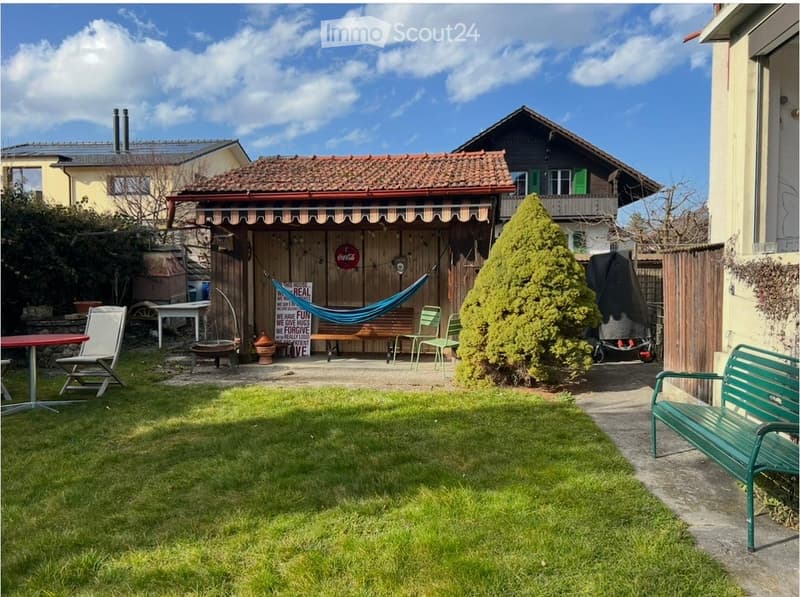Garden with a grassy lawn, a small wooden shed, a hammock, outdoor furniture including a red table, white folding chair, green chairs, and a green bench. There is also a fire pit and a large bush.