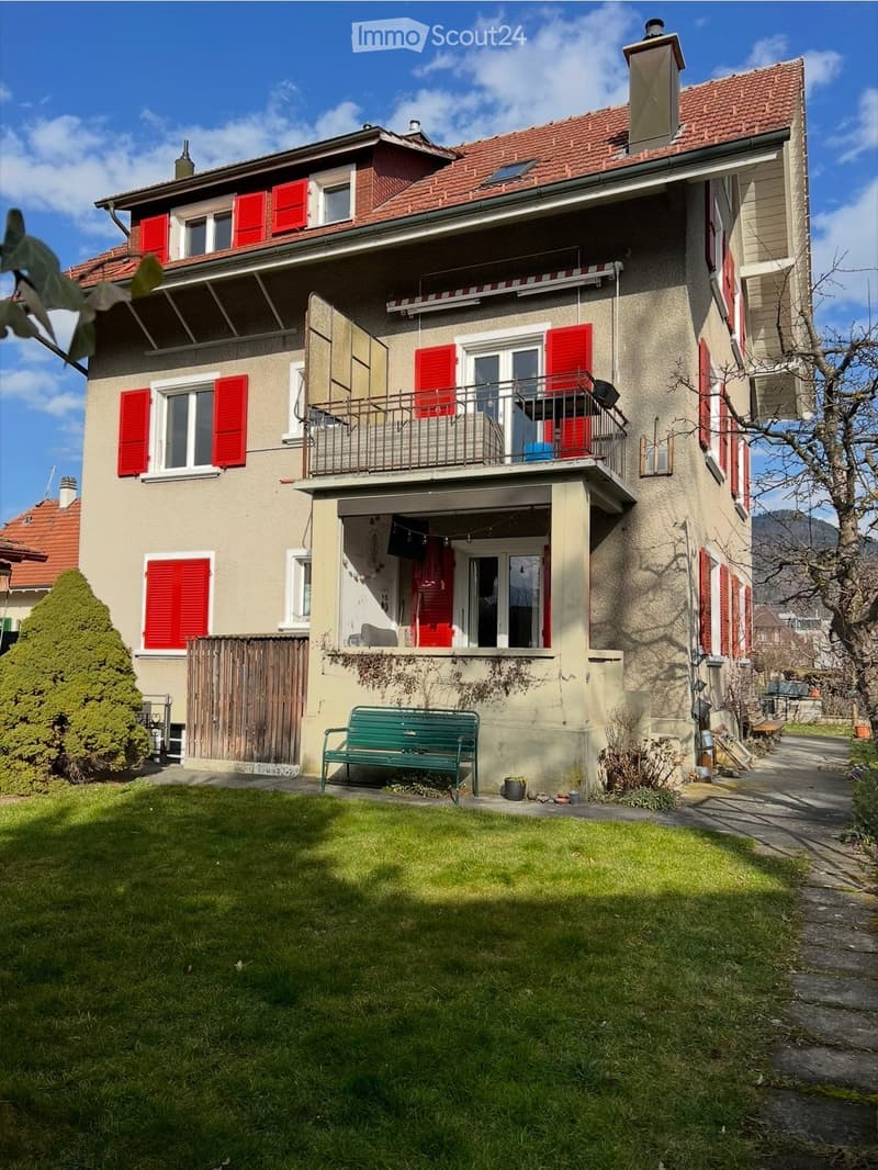 Two-story house with red shutters, beige walls, and a red-tiled roof. Features include a balcony, a green bench in the front yard, and a small garden area.