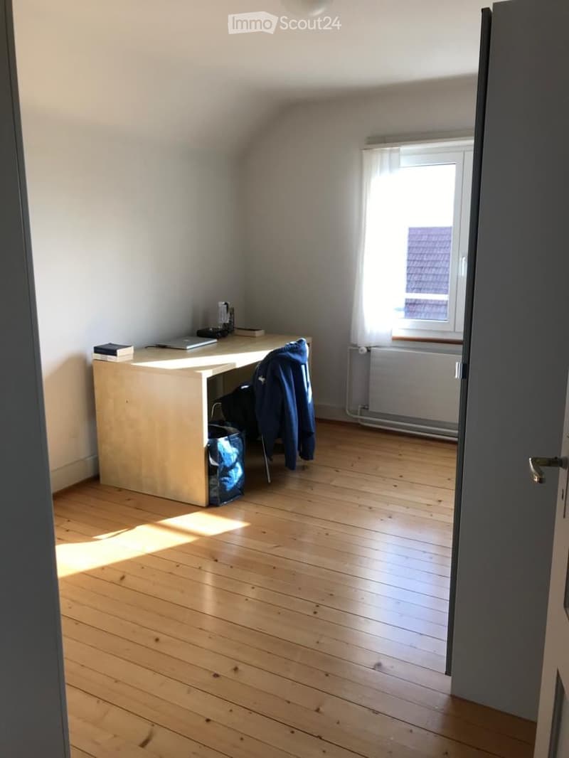 Bright room with wooden flooring, a desk with a laptop and books, a chair, and a window with a curtain.