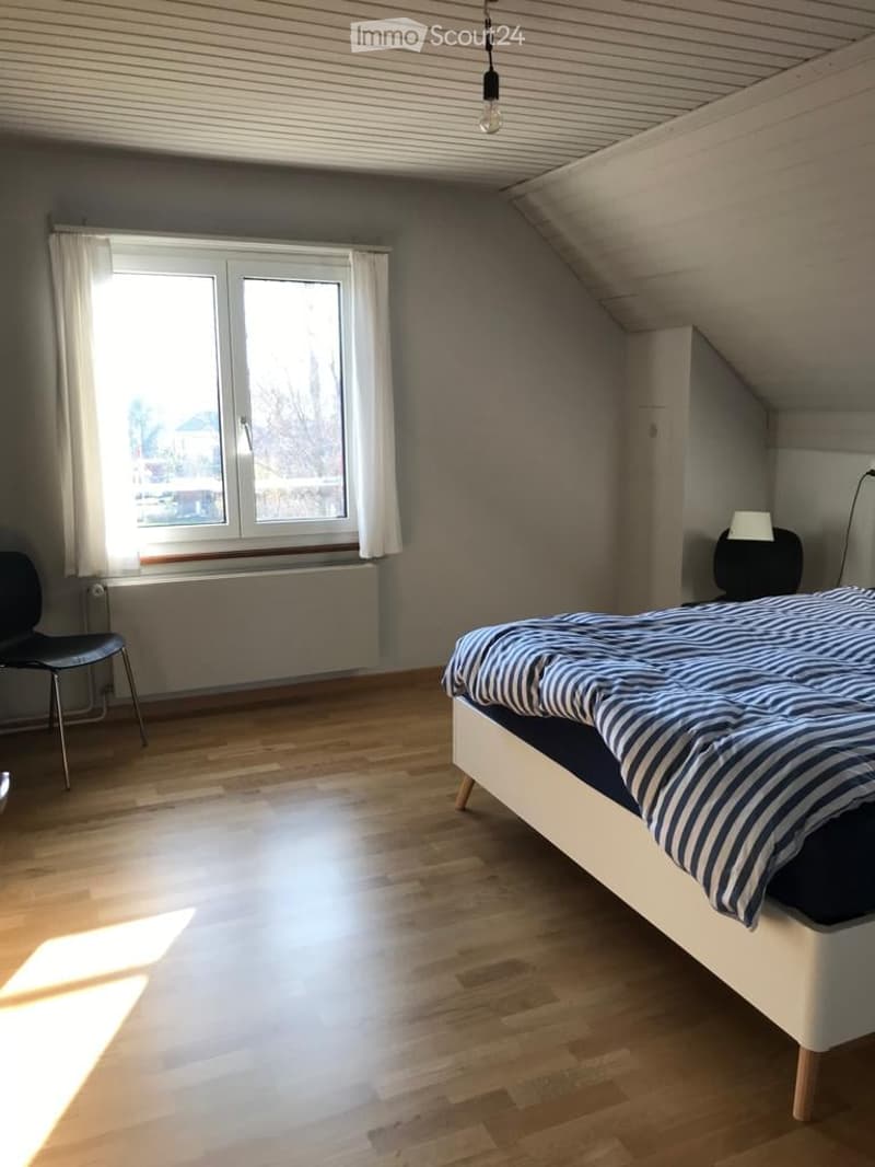 Bright bedroom with wooden flooring, large window with white curtains, bed with striped bedding, black chair, and a small table lamp.