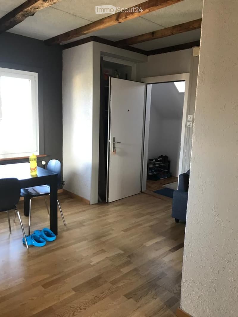 Bright room with wooden flooring, a small table with chairs, a bottle on the table, and a closet with an open door revealing some items inside.
