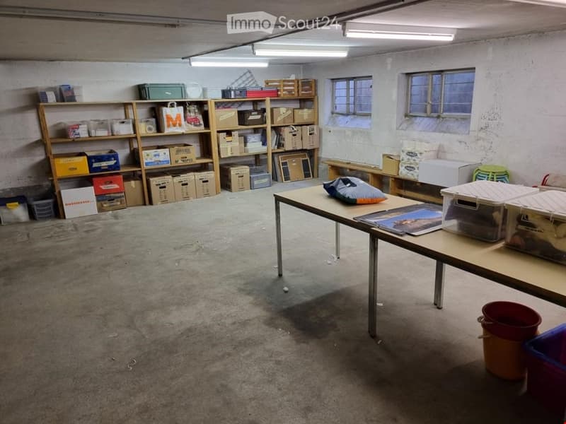 The room is a storage or workspace with concrete floors, exposed pipes, and fluorescent lighting. It features a large wooden table with a pillow and some items on it, shelves filled with boxes and storage items, and a few stools.