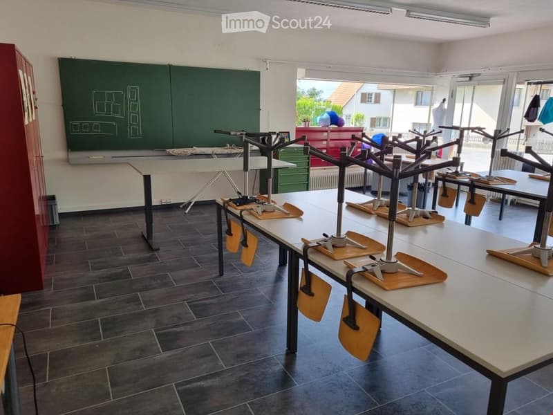 Classroom with long tables, chairs, a blackboard, and a cabinet. Large windows provide natural light.