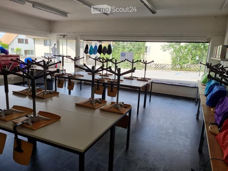 A spacious room with multiple tables and chairs arranged for a workshop or studio setting. Mannequins and racks for clothing or accessories are visible, along with various tools and materials.