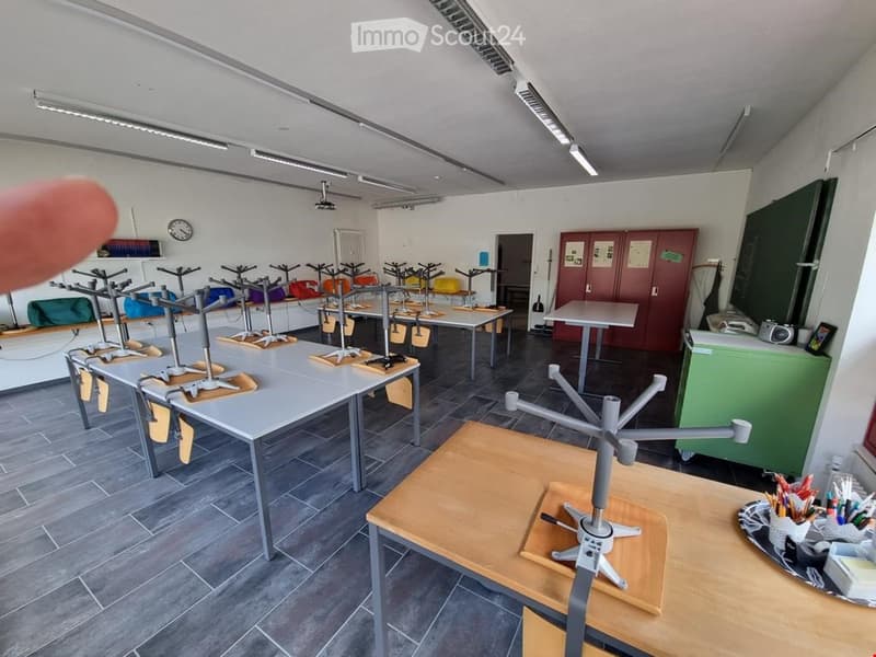 Classroom or workshop space with multiple tables, chairs, and cabinets. The room features a tiled floor, white walls, and a clock on the wall.