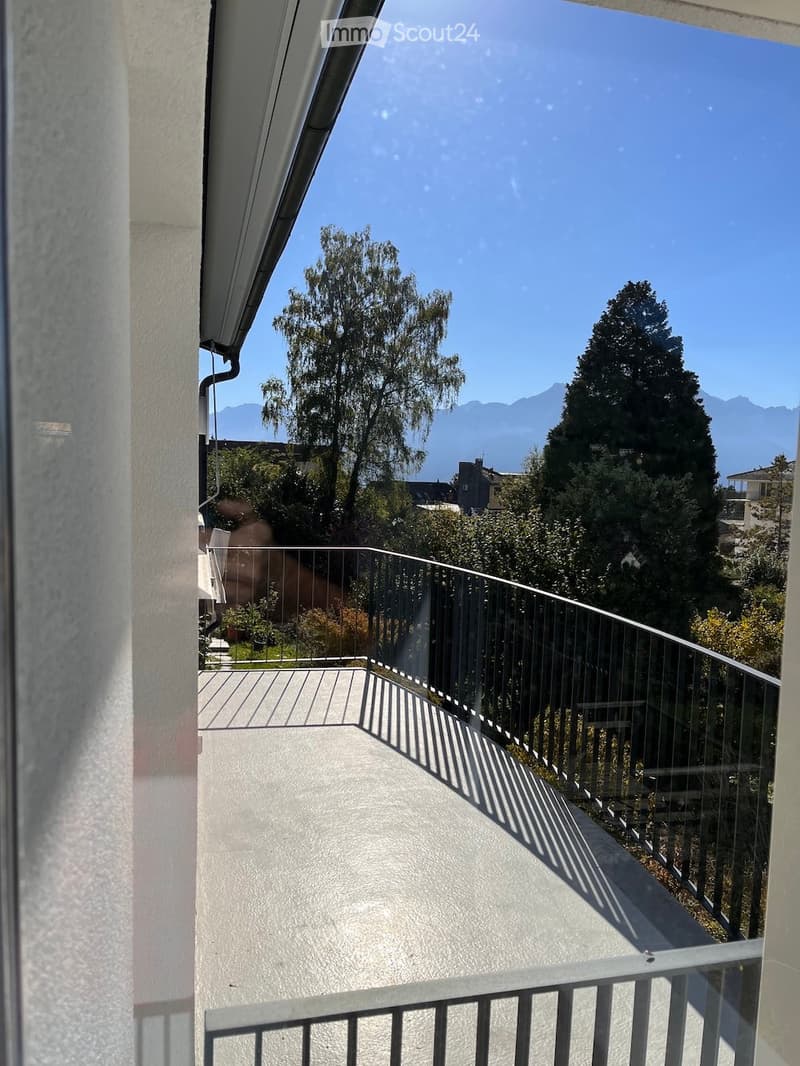 Balcony with a modern metal railing, overlooking a lush green area with trees and distant mountains under a clear blue sky.