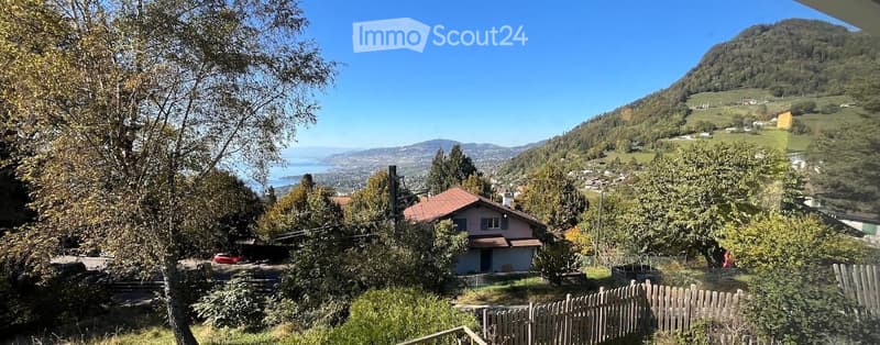 A scenic outdoor view from a balcony or terrace showing a house surrounded by trees, a wooden fence, and a panoramic view of a valley with mountains and a lake in the distance.
