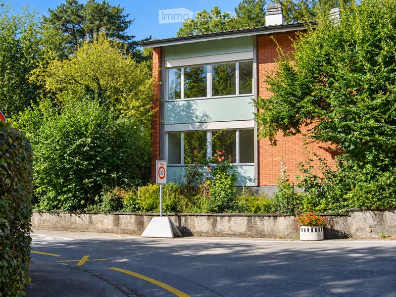 Two-story house with red brick and light green siding, surrounded by lush greenery and trees. Features large windows and a small balcony area with flowers.