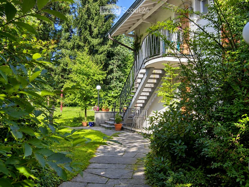 A well-maintained garden area with a stone pathway leading to a house entrance, featuring potted plants, a small seating area, and lush greenery.
