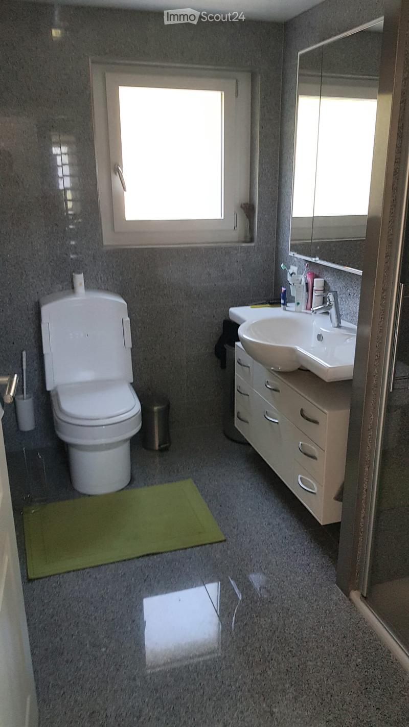 Modern bathroom with grey walls, a white toilet, a sink with a mirror, a cabinet, and a green bath mat on the floor.