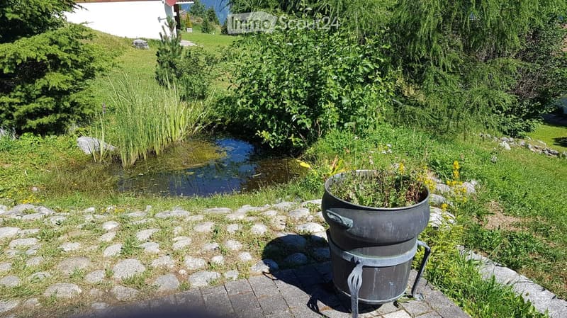 A serene garden area with a small pond surrounded by lush greenery, a stone pathway, and a large metal container with plants on the right side.